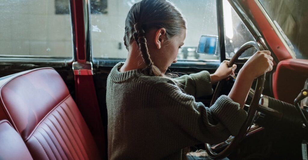 A girl with pigtails sits in a vintage car, playfully holding the steering wheel.